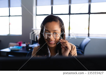 African American woman sitting at office desk wearing headset and glasses, working with notebook African American woman sitting at office desk wearing headset and glasses, working with notebook 135355427