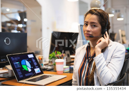 Woman in blazer handling calls with headset using laptop showing charts in office, copy space 135355434