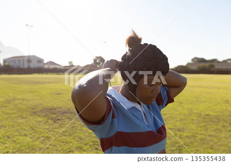 African American teenage girl wearing rugby jersey adjusting padded headguard on grass field 135355438