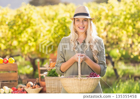Blonde woman wearing straw hat and smiling in vineyard at table holding basket of red grapes Blonde woman wearing straw hat and smiling in vineyard at table holding basket of red grapes 135355458