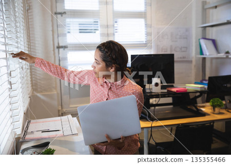 African American woman adjusting blinds at design studio holding laptop and clipboard, copy space 135355466