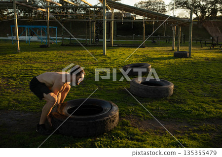 Man wearing black shorts squatting on field flipping tractor tire near obstacle frame, copy space Man wearing black shorts squatting on field flipping tractor tire near obstacle frame, copy space 135355479