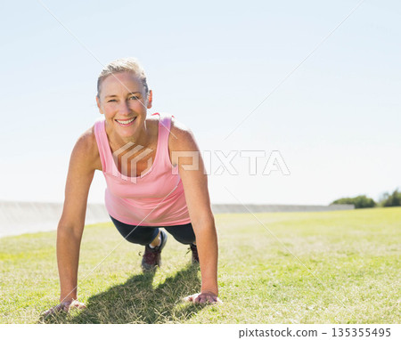 Mature adult woman holding plank on grassy field at park showing athletic shoes and leggings 135355495