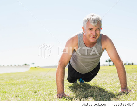 Senior male performing push-ups on grass field wearing grey tank top and blue-accented sneakers 135355497