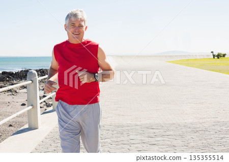 Senior man jogging along paved seaside promenade checking wristwatch near metal railing, copy space 135355514