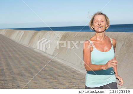 Woman jogging along seafront promenade beside concrete seawall under clear blue sky, copy space Woman jogging along seafront promenade beside concrete seawall under clear blue sky, copy space 135355519