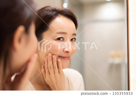 一位日本女性正在鏡子前檢查皮膚。圖片展示了護膚和美容流程。 135355933