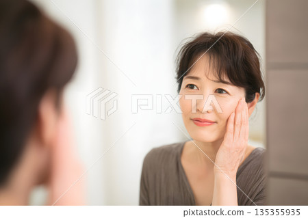 一位日本女性正在鏡子前檢查皮膚。圖片展示了護膚和美容流程。 135355935
