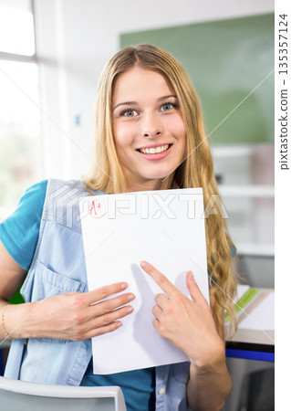 Female teenage student sitting at desk under chalkboard holding graded sheet showing red plus mark 135357124