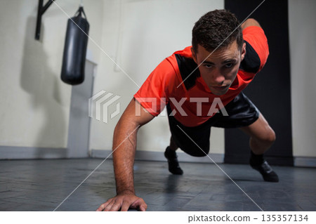 Male in red shirt performing one-arm pushup on rubber gym floor with punching bag, wall pad 135357134