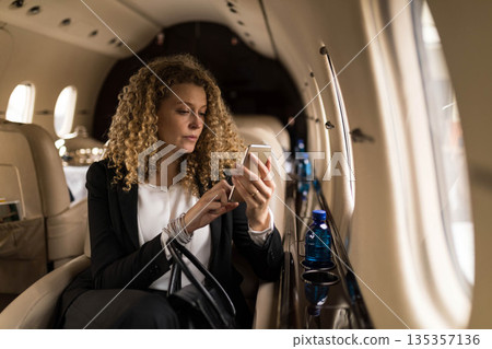 Businesswoman tapping smartphone screen in private jet cabin with blue water bottle 135357136
