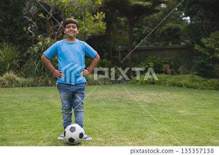 Boy standing with hands on hips wearing blue t-shirt on green backyard lawn with soccer ball 135357138