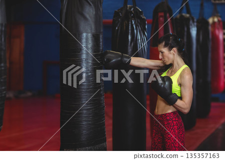 Mid adult woman punching heavy bag at boxing gym in neon sportswear and gloves, copy space Mid adult woman punching heavy bag at boxing gym in neon sportswear and gloves, copy space 135357163