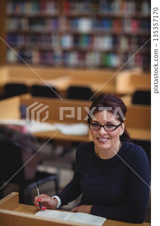 Mid adult woman sitting at library carrel wearing eyeglasses holding pen over open book, copy space 135357170