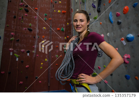 Female rock climber holding coiled rope over shoulder in climbing gym wearing harness, copy space 135357179