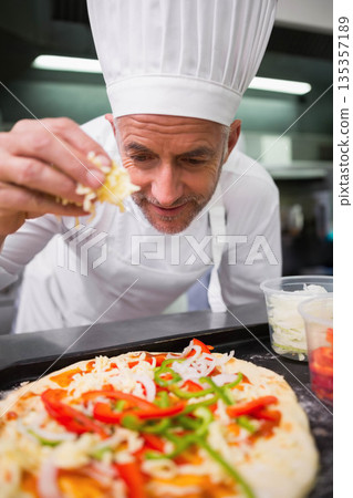 Male chef wearing white toque jacket sprinkling shredded cheese onto pizza crust at kitchen counter 135357189