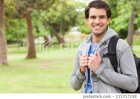 Man standing smiling in park wearing grey jacket blue shirt carrying black backpack, copy space 135357198