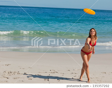 Woman in red bikini standing on sandy beach tossing yellow flying disc, clear sky, copy space Woman in red bikini standing on sandy beach tossing yellow flying disc, clear sky, copy space 135357202