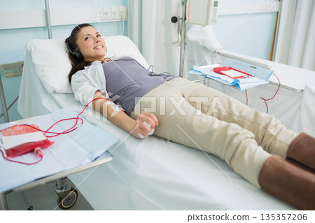 Female donor reclining on hospital bed with blood bag, needle and headphones in donation room Female donor reclining on hospital bed with blood bag, needle and headphones in donation room 135357206