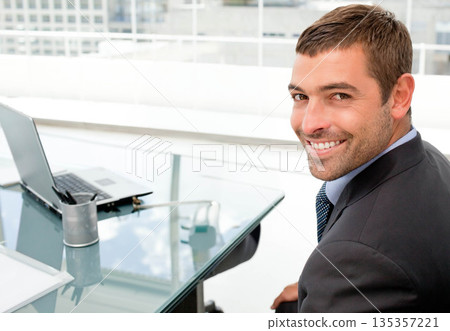Man wearing suit smiling while working at glass-top desk by window with laptop, papers, copy space 135357221