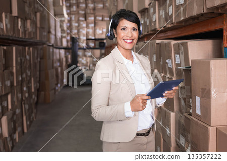 Asian woman wearing beige business suit inspecting racks in warehouse holding tablet and headset 135357222