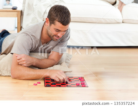 Man in thirties lying on floor cushion at home playing checkers on red and black board 135357224