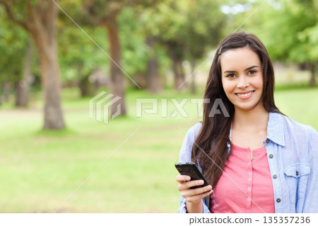 Teenage woman standing in grassy park holding smartphone while smiling at camera Teenage woman standing in grassy park holding smartphone while smiling at camera 135357236