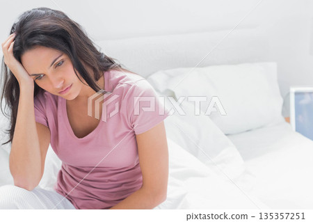 Woman sitting on unmade bed wearing pink shirt and striped pajama pants leaning forward in bedroom 135357251