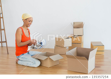 African American woman kneeling in empty room packing moving boxes with newspaper, copy space 135357254