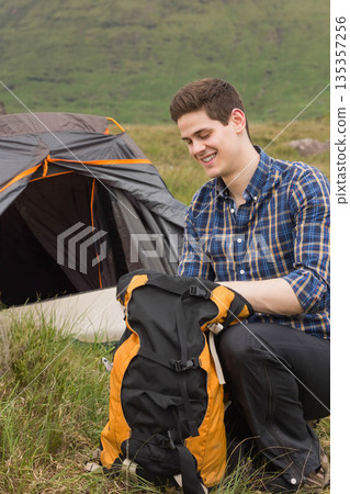 Man kneeling next to gray dome tent on grassy hillside, adjusting orange black backpack, copy space 135357256