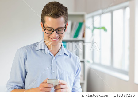 Businessman standing in office holding smartphone wearing glasses by bookshelf with books and plant 135357259