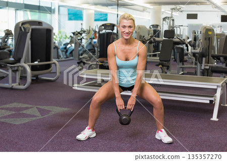 Fit woman Holding black kettlebell in both hands on purple carpet at gym wearing sportswear Fit woman Holding black kettlebell in both hands on purple carpet at gym wearing sportswear 135357270