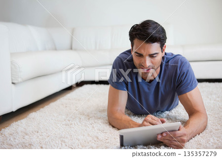 Mid-adult Hispanic male lying on white shag carpet at home holding tablet near white leather sofa Mid-adult Hispanic male lying on white shag carpet at home holding tablet near white leather sofa 135357279