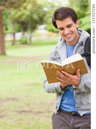 Male student standing on grassy lawn in park reading hardcover book with backpack, copy space 135357284