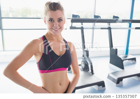 Female athlete posing with hands on hips in gym with stationary bike, step platforms, copy space Female athlete posing with hands on hips in gym with stationary bike, step platforms, copy space 135357307