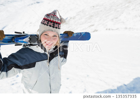 Standing woman on snow covered slope wearing winter jacket, knit hat holding blue skis, copy space 135357335