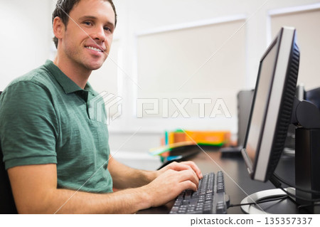 Man in his thirties smiling and typing on keyboard beside dual monitors in office, copy space 135357337