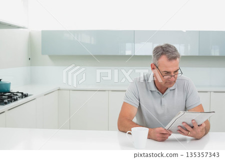 Man reading newspaper while sitting at kitchen island with coffee mug and teal pot, copy space 135357343
