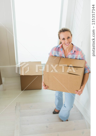 Female carrying cardboard box climbing staircase at home near frosted window, copy space 135357344