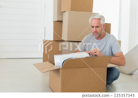 Man kneeling on floor in bright home unwrapping bubble-wrapped item from cardboard box, copy space 135357345