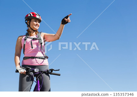 Mid adult female cyclist straddling purple bicycle on trail pointing ahead with helmet, copy space 135357346