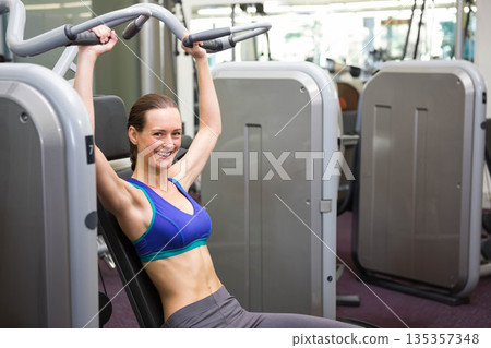 Adult woman wearing sports bra pushing chest press machine handles at gym by mirrors, copy space 135357348