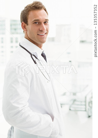 Male doctor wearing white lab coat and tie, standing near windows in clinic with stethoscope Male doctor wearing white lab coat and tie, standing near windows in clinic with stethoscope 135357352