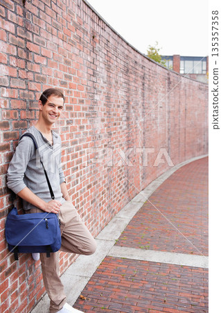 Man leaning on brick-paved walkway beside red brick wall holding blue messenger bag, copy space Man leaning on brick-paved walkway beside red brick wall holding blue messenger bag, copy space 135357358