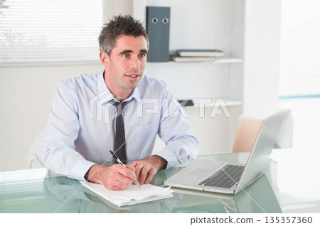 Man in dress shirt and tie writing on paperwork at glass-top desk in office with laptop 135357360