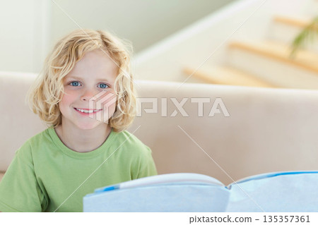 School-age boy wearing green T-shirt holding open book and smiling at camera on couch at home 135357361