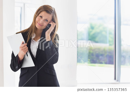 Woman standing in bright modern office holding clipboard and speaking into smartphone, copy space Woman standing in bright modern office holding clipboard and speaking into smartphone, copy space 135357365