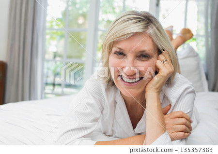 Senior woman wearing pajamas lying on white sheets in bedroom smiling at camera by sheer curtains 135357383