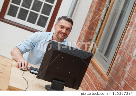 Mid adult man sitting at wooden office desk smiling while using desktop monitor and wired mouse Mid adult man sitting at wooden office desk smiling while using desktop monitor and wired mouse 135357384