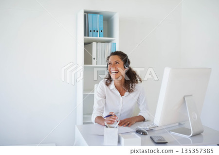 Woman sitting at office desk wearing headset, smiling and writing paperwork by monitor and binders Woman sitting at office desk wearing headset, smiling and writing paperwork by monitor and binders 135357385